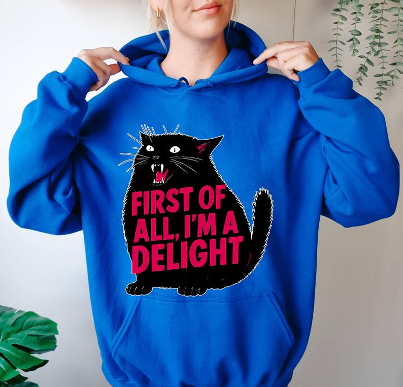 Op de afbeelding: Koningsblauwe hoodie met een zwarte kattenafbeelding. De kat heeft een open mond en de tekst "FIRST OF ALL, I'M A DELIGHT" in roze. De hoodie heeft een capuchon met trekkoord.