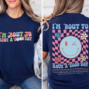 Puede incluir: Camiseta azul marino con el texto "I'M 'BOUT TO HAVE A GOOD DAY" en una fuente retro colorida. La parte trasera presenta una carita sonriente gui&ntilde;ando un ojo sobre un fondo a cuadros rosa y azul, con flores y texto.