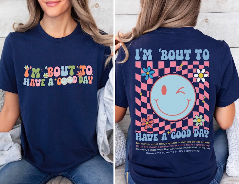 Puede incluir: Camiseta azul marino con el texto "I'M 'BOUT TO HAVE A GOOD DAY" en una fuente retro colorida. La parte trasera presenta una carita sonriente gui&ntilde;ando un ojo sobre un fondo a cuadros rosa y azul, con flores y texto.