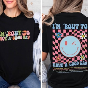 Puede incluir: Camiseta negra con la frase "I'M 'BOUT TO HAVE A GOOD DAY" en letras coloridas de estilo retro. La parte trasera de la camiseta presenta una carita sonriente gui&ntilde;ando un ojo sobre un fondo a cuadros rosa y negro, con texto y flores adicionales.