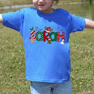 Op de afbeelding: Een blauw T-shirt met een kerstthema ontwerp met de naam "Sarah" in kleurrijke letters met feestelijke versieringen.