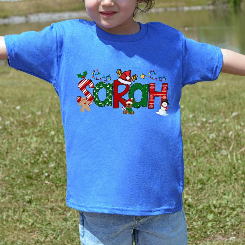 Op de afbeelding: Een blauw T-shirt met een kerstthema ontwerp met de naam "Sarah" in kleurrijke letters met feestelijke versieringen.