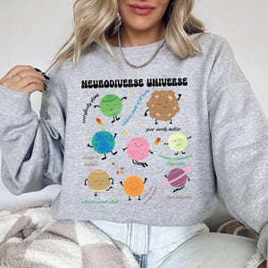 K&ouml;nnte beinhalten: Grauer Pullover mit einem Grafikdruck eines bunten Sonnensystems mit Planeten und Sternen. Der Text "Neurodiverse Universe" ist auf der Vorderseite des Pullovers aufgedruckt. Die Planeten haben Gesichter und sind mit S&auml;tzen wie "Jeder gl&auml;nzt", "Feiert die K&ouml;pfe aller Arten", "Deine Worte z&auml;hlen", "&Auml;ndere dich, um einzuschlie&szlig;en", "Jeder kommuniziert anders", "Denke anders", "Bilde, akzeptiere, passe dich an", "Wir sind das Licht" beschriftet.