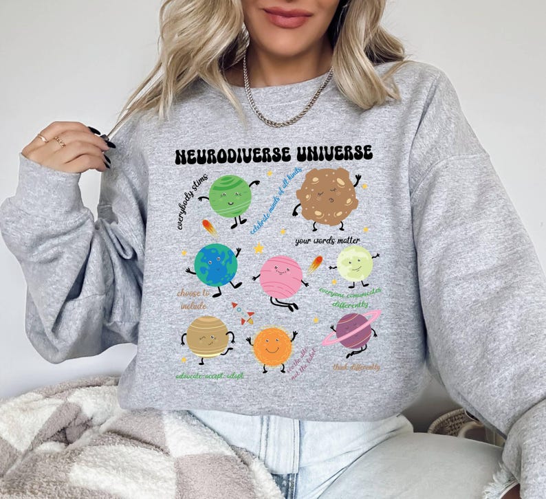K&ouml;nnte beinhalten: Grauer Pullover mit einem Grafikdruck eines bunten Sonnensystems mit Planeten und Sternen. Der Text "Neurodiverse Universe" ist auf der Vorderseite des Pullovers aufgedruckt. Die Planeten haben Gesichter und sind mit S&auml;tzen wie "Jeder gl&auml;nzt", "Feiert die K&ouml;pfe aller Arten", "Deine Worte z&auml;hlen", "&Auml;ndere dich, um einzuschlie&szlig;en", "Jeder kommuniziert anders", "Denke anders", "Bilde, akzeptiere, passe dich an", "Wir sind das Licht" beschriftet.