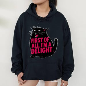 Op de afbeelding: Zwarte hoodie met een cartoon zwarte kat en de roze tekst "FIRST OF ALL, I'M A DELIGHT". De kat heeft witte snorharen en een open mond. De hoodie heeft een trekkoord en een voorzak.
