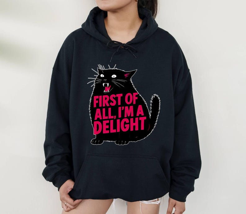 Op de afbeelding: Zwarte hoodie met een cartoon zwarte kat en de roze tekst "FIRST OF ALL, I'M A DELIGHT". De kat heeft witte snorharen en een open mond. De hoodie heeft een trekkoord en een voorzak.