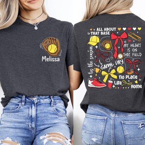 May include: Two gray t-shirts with a baseball theme. One shirt has a baseball glove and ball with the name "Melissa" on it. The other shirt has a variety of baseball-themed graphics, including a baseball glove, a baseball, a trophy, a bow, and the text "All About That Base", "My Heart Is On That Field", "Game Day", "No Place Like Home", and "Tis The Season".
