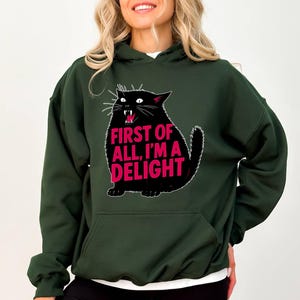 Op de afbeelding: Donkergroene hoodie met een zwarte kattenafbeelding en de roze tekst "FIRST OF ALL, I'M A DELIGHT". De kat heeft een open mond en witte snorharen. De hoodie heeft een voorzak en een capuchon.