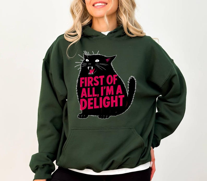 Op de afbeelding: Donkergroene hoodie met een zwarte kattenafbeelding en de roze tekst "FIRST OF ALL, I'M A DELIGHT". De kat heeft een open mond en witte snorharen. De hoodie heeft een voorzak en een capuchon.