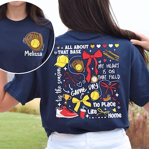 May include: Navy blue t-shirt with a baseball-themed design on the back. The design includes a baseball glove, a baseball, a trophy, a bow, a baseball cap, bats, and the text "tis the season", "ALL ABOUT THAT BASE", "GAME DAY", "MY HEART IS ON THAT FIELD", "no Place Like Home".