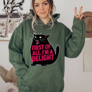 Op de afbeelding: Een olijfgroene hoodie met een zwarte kattenafbeelding en de roze tekst "FIRST OF ALL, I'M A DELIGHT". De kat heeft witte snorharen en een rode mond. De hoodie heeft een capuchon met trekkoord en een voorzak.