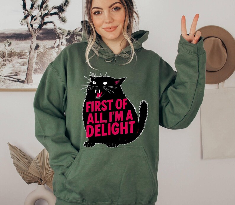 Op de afbeelding: Een olijfgroene hoodie met een zwarte kattenafbeelding en de roze tekst "FIRST OF ALL, I'M A DELIGHT". De kat heeft witte snorharen en een rode mond. De hoodie heeft een capuchon met trekkoord en een voorzak.
