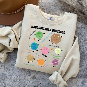 K&ouml;nnte beinhalten: Ein hellbeiger Pullover mit einer Grafik von Planeten und dem Text "Neurodiverse Universe" mit S&auml;tzen wie "Jeder stimt", "Feiert die K&ouml;pfe aller Arten", "Eure Worte z&auml;hlen", "Jeder kommuniziert anders", "W&auml;hlt das Einschlie&szlig;en", "Vertreten-Akzeptieren-Anpassen", "Der Himmel ist nicht die Grenze", "Denkt &uuml;ber den Tellerrand hinaus".