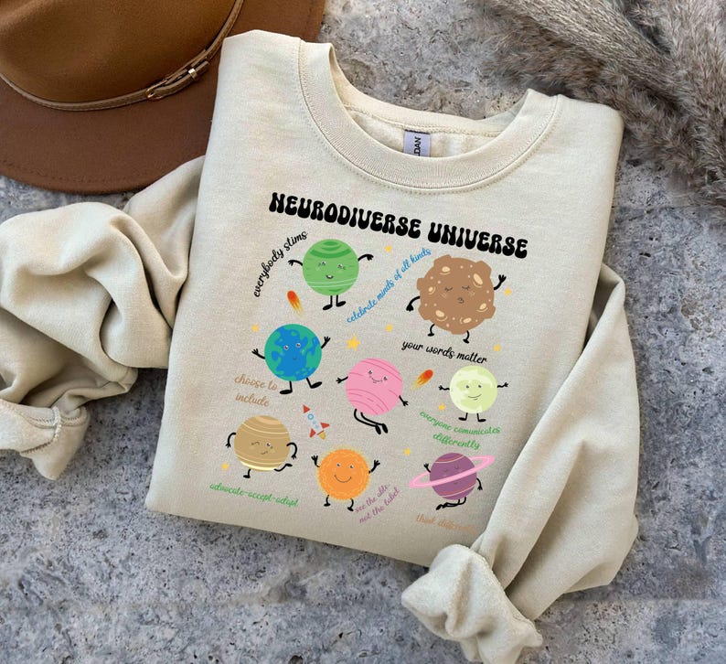 K&ouml;nnte beinhalten: Ein hellbeiger Pullover mit einer Grafik von Planeten und dem Text "Neurodiverse Universe" mit S&auml;tzen wie "Jeder stimt", "Feiert die K&ouml;pfe aller Arten", "Eure Worte z&auml;hlen", "Jeder kommuniziert anders", "W&auml;hlt das Einschlie&szlig;en", "Vertreten-Akzeptieren-Anpassen", "Der Himmel ist nicht die Grenze", "Denkt &uuml;ber den Tellerrand hinaus".