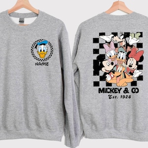 Puede incluir: Dos sudaderas grises con diferentes dise&ntilde;os de personajes de Disney. Una sudadera presenta una imagen de dibujos animados de Donald Duck con el texto "Name". La otra sudadera presenta un grupo de personajes de Disney, incluidos Mickey Mouse, Minnie Mouse, Donald Duck, Goofy y Pluto. El texto "Mickey & Co. Est. 1928" est&aacute; impreso debajo de los personajes.