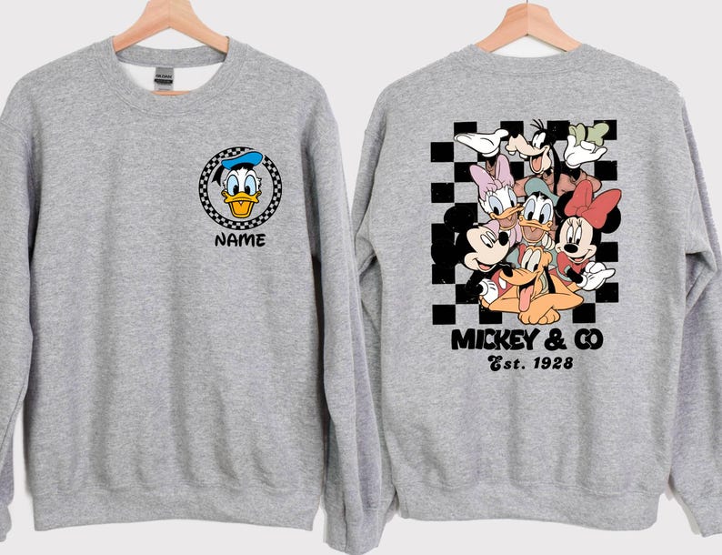 Puede incluir: Dos sudaderas grises con diferentes dise&ntilde;os de personajes de Disney. Una sudadera presenta una imagen de dibujos animados de Donald Duck con el texto "Name". La otra sudadera presenta un grupo de personajes de Disney, incluidos Mickey Mouse, Minnie Mouse, Donald Duck, Goofy y Pluto. El texto "Mickey & Co. Est. 1928" est&aacute; impreso debajo de los personajes.