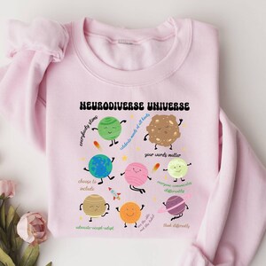 K&ouml;nnte beinhalten: Rosafarbener Pullover mit einem grafischen Design von Planeten und Sternen mit dem Text "Neurodiverse Universe" und S&auml;tzen wie "everybody slims", "celebrate minds of all kinds", "your words matter", "choose to include", "advocate-accept-adapt", "everyone communicates differently", "we are the tribe, not the label", and "think differently".