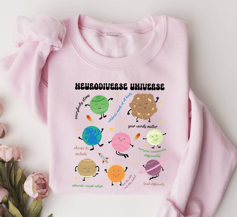 K&ouml;nnte beinhalten: Rosafarbener Pullover mit einem grafischen Design von Planeten und Sternen mit dem Text "Neurodiverse Universe" und S&auml;tzen wie "everybody slims", "celebrate minds of all kinds", "your words matter", "choose to include", "advocate-accept-adapt", "everyone communicates differently", "we are the tribe, not the label", and "think differently".