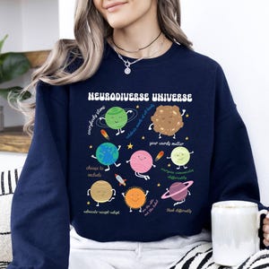 K&ouml;nnte beinhalten: Ein marineblauer Sweatshirt mit einer Grafik von Planeten und dem Text "Neurodiverse Universe". Die Planeten haben Gesichter und jeder Planet hat eine andere Botschaft. Die Botschaften lauten: "Jeder stimt", "Feiert Worte und alles, was sie beinhalten", "Deine Worte z&auml;hlen", "Jeder kommuniziert anders", "W&auml;hlt, einzuschlie&szlig;en", "Bef&uuml;rworten-Akzeptieren-Anpassen", "Seht das Kleine, nicht das Etikett", und "Denkt anders".