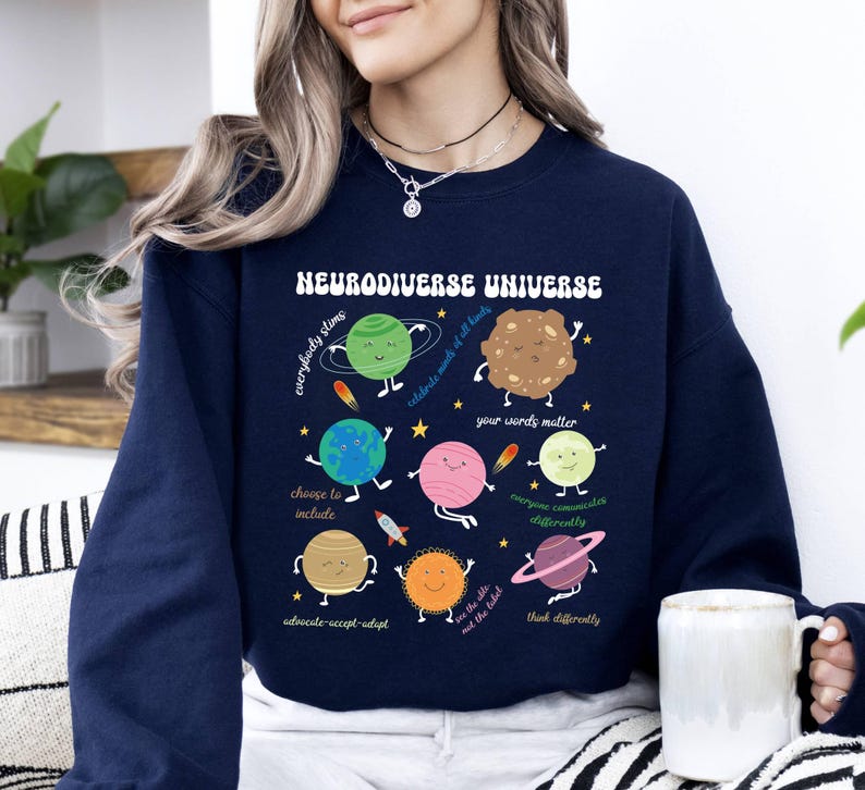 K&ouml;nnte beinhalten: Ein marineblauer Sweatshirt mit einer Grafik von Planeten und dem Text "Neurodiverse Universe". Die Planeten haben Gesichter und jeder Planet hat eine andere Botschaft. Die Botschaften lauten: "Jeder stimt", "Feiert Worte und alles, was sie beinhalten", "Deine Worte z&auml;hlen", "Jeder kommuniziert anders", "W&auml;hlt, einzuschlie&szlig;en", "Bef&uuml;rworten-Akzeptieren-Anpassen", "Seht das Kleine, nicht das Etikett", und "Denkt anders".