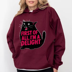 Op de afbeelding: Bordeaux hoodie met een zwarte kat illustratie en de roze tekst "FIRST OF ALL, I'M A DELIGHT". De kat heeft witte snorharen en een rode mond. De hoodie heeft een voorzak en een capuchon.