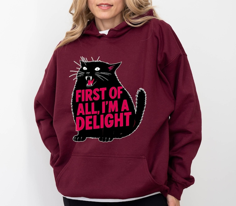 Op de afbeelding: Bordeaux hoodie met een zwarte kat illustratie en de roze tekst "FIRST OF ALL, I'M A DELIGHT". De kat heeft witte snorharen en een rode mond. De hoodie heeft een voorzak en een capuchon.