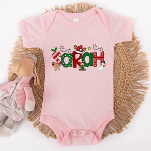 Op de afbeelding: Roze baby-romper met een kerstthema met de naam "Sarah" in feestelijke letters met kerstversiering.