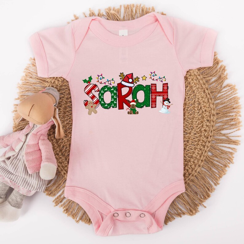Op de afbeelding: Roze baby-romper met een kerstthema met de naam "Sarah" in feestelijke letters met kerstversiering.