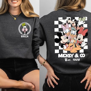 Puede incluir: Una sudadera gris con un patr&oacute;n a cuadros y un gr&aacute;fico de Mickey Mouse y sus amigos. El texto "Mickey & Co. Est. 1928" est&aacute; impreso debajo del gr&aacute;fico. La sudadera tiene un bolsillo en el lado izquierdo con un gr&aacute;fico de Goofy y el nombre "Erica" impreso debajo.