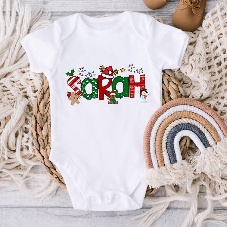 Op de afbeelding: Witte baby romper met een kerstthema ontwerp met de naam "SARAH" in kleurrijke letters met feestelijke decoraties zoals snoepstokken, lichtjes en een sneeuwpop.