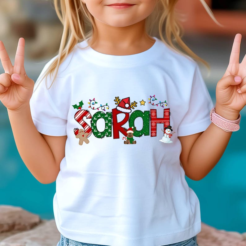 Op de afbeelding: Wit T-shirt met een kerstthema ontwerp met de naam "Sarah" in kleurrijke letters met feestelijke decoraties zoals snoepstokken, lichtjes en een sneeuwpop.
