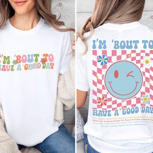 Puede incluir: Camiseta blanca con el texto "I'M 'BOUT TO HAVE A GOOD DAY" en letras coloridas de estilo retro. La parte trasera de la camiseta presenta una carita sonriente gui&ntilde;ando un ojo y la misma frase, sobre un patr&oacute;n de cuadros rosas y azules.