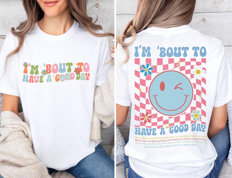 Puede incluir: Camiseta blanca con el texto "I'M 'BOUT TO HAVE A GOOD DAY" en letras coloridas de estilo retro. La parte trasera de la camiseta presenta una carita sonriente gui&ntilde;ando un ojo y la misma frase, sobre un patr&oacute;n de cuadros rosas y azules.