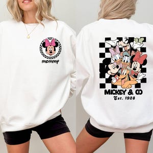 Puede incluir: Sudadera blanca con un patr&oacute;n de cuadros blanco y negro y un grupo de personajes de Disney que incluyen a Mickey Mouse, Minnie Mouse, Donald Duck, Goofy y Pluto. El texto "Mickey & Co. Est. 1928" est&aacute; impreso debajo de los personajes. La parte delantera de la sudadera tiene una imagen en blanco y negro de Minnie Mouse con la palabra "mam&aacute;" impresa debajo.