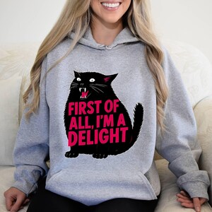 Op de afbeelding: Een grijze hoodie met een zwarte kattenafbeelding en de roze tekst "FIRST OF ALL, I'M A DELIGHT". De kat heeft een open mond met zichtbare hoektanden. De hoodie heeft een capuchon met trekkoord en lange mouwen.