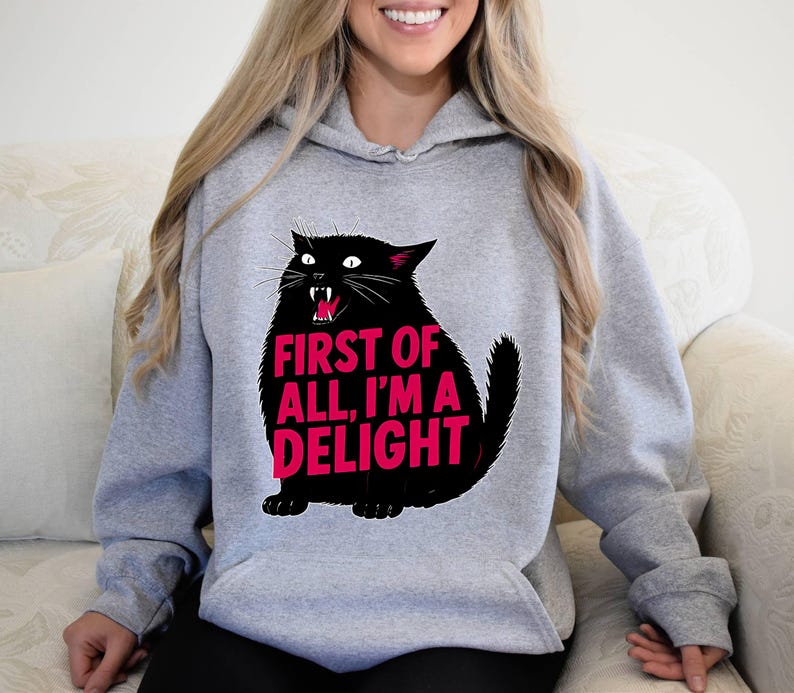Op de afbeelding: Een grijze hoodie met een zwarte kattenafbeelding en de roze tekst "FIRST OF ALL, I'M A DELIGHT". De kat heeft een open mond met zichtbare hoektanden. De hoodie heeft een capuchon met trekkoord en lange mouwen.