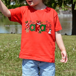 Op de afbeelding: Rood T-shirt met een kerstthema ontwerp met de naam "Sarah" in groene en rode letters met feestelijke versieringen.
