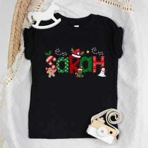 Op de afbeelding: Zwart kerst-thema T-shirt met de naam "Sarah" gespeld in feestelijke letters met kerstversiering.