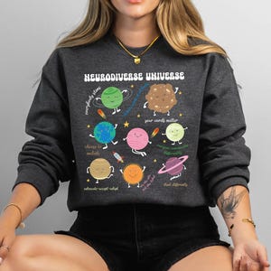 K&ouml;nnte beinhalten: Ein dunkelgraues Sweatshirt mit einem grafischen Aufdruck eines Sonnensystems mit Planeten, die Gesichter und Text haben. Der Text auf den Planeten lautet "Jeder strahlt", "W&auml;hle, einzuschlie&szlig;en", "Deine Worte z&auml;hlen", "Jeder kommuniziert anders", "Anders denken", "Vertreten-Akzeptieren-Anpassen", und "Wir sind die Wildnis". Der Text oben auf der Grafik lautet "Neurodiverses Universum".