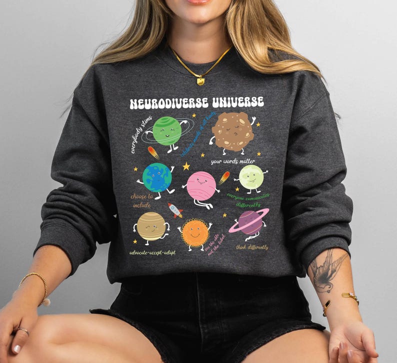 K&ouml;nnte beinhalten: Ein dunkelgraues Sweatshirt mit einem grafischen Aufdruck eines Sonnensystems mit Planeten, die Gesichter und Text haben. Der Text auf den Planeten lautet "Jeder strahlt", "W&auml;hle, einzuschlie&szlig;en", "Deine Worte z&auml;hlen", "Jeder kommuniziert anders", "Anders denken", "Vertreten-Akzeptieren-Anpassen", und "Wir sind die Wildnis". Der Text oben auf der Grafik lautet "Neurodiverses Universum".