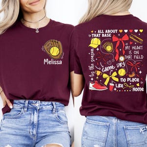 May include: Two maroon t-shirts with a softball theme. The front shirt has a baseball glove with a ball and the name "Melissa". The back shirt has a variety of softball-related graphics and the text "tis the season", "ALL ABOUT THAT BASE", "MY HEART IS ON THAT FIELD", "GAME DAY", "No Place Like Home".