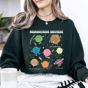 K&ouml;nnte beinhalten: Gr&uuml;nes Sweatshirt mit einer Grafik eines Sonnensystems mit Planeten und dem Text "Neurodiverse Universe" mit S&auml;tzen wie "Jeder stimt", "W&auml;hle, um einzuschlie&szlig;en", "Deine Worte z&auml;hlen", "Jeder kommuniziert anders", "Sieh die Welt, nicht das Etikett", und "Denke anders".