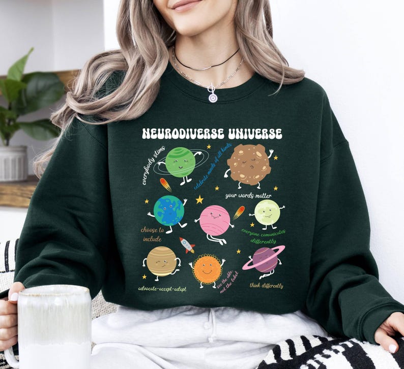K&ouml;nnte beinhalten: Gr&uuml;nes Sweatshirt mit einer Grafik eines Sonnensystems mit Planeten und dem Text "Neurodiverse Universe" mit S&auml;tzen wie "Jeder stimt", "W&auml;hle, um einzuschlie&szlig;en", "Deine Worte z&auml;hlen", "Jeder kommuniziert anders", "Sieh die Welt, nicht das Etikett", und "Denke anders".