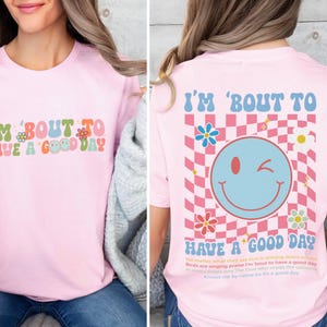 Puede incluir: Camiseta rosa claro con la frase "I'M 'BOUT TO HAVE A GOOD DAY" en letras retro coloridas. La parte trasera de la camiseta presenta una carita sonriente gui&ntilde;ando un ojo sobre un fondo a cuadros rosa y blanco, con flores y texto.