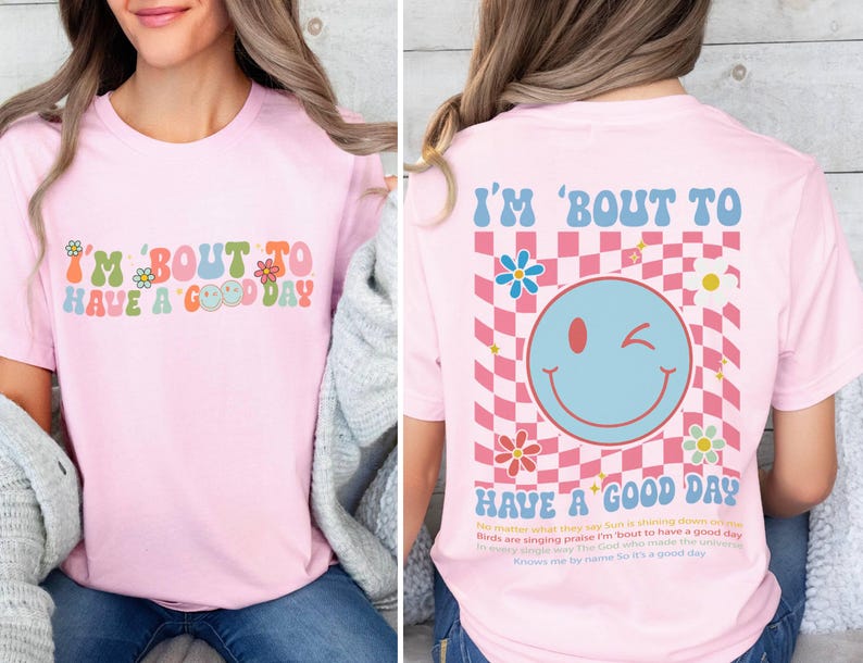 Puede incluir: Camiseta rosa claro con la frase "I'M 'BOUT TO HAVE A GOOD DAY" en letras retro coloridas. La parte trasera de la camiseta presenta una carita sonriente gui&ntilde;ando un ojo sobre un fondo a cuadros rosa y blanco, con flores y texto.