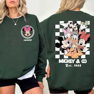 Puede incluir: Sudadera verde con un gr&aacute;fico de Mickey Mouse y sus amigos en la espalda. El gr&aacute;fico presenta a Mickey Mouse, Minnie Mouse, Donald Duck, Goofy, Pluto y un fondo a cuadros. El texto "Mickey & Co. Est. 1928" est&aacute; impreso debajo del gr&aacute;fico. La parte delantera de la sudadera presenta un peque&ntilde;o gr&aacute;fico de Minnie Mouse con el texto "mommy" impreso debajo.