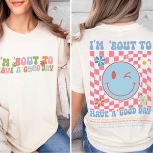 Puede incluir: Camiseta color crema con el texto "I'M 'BOUT TO HAVE A GOOD DAY" en letras coloridas de estilo retro. La parte trasera presenta una gran carita sonriente gui&ntilde;ando un ojo sobre un fondo a cuadros rosa y blanco, con flores y texto.