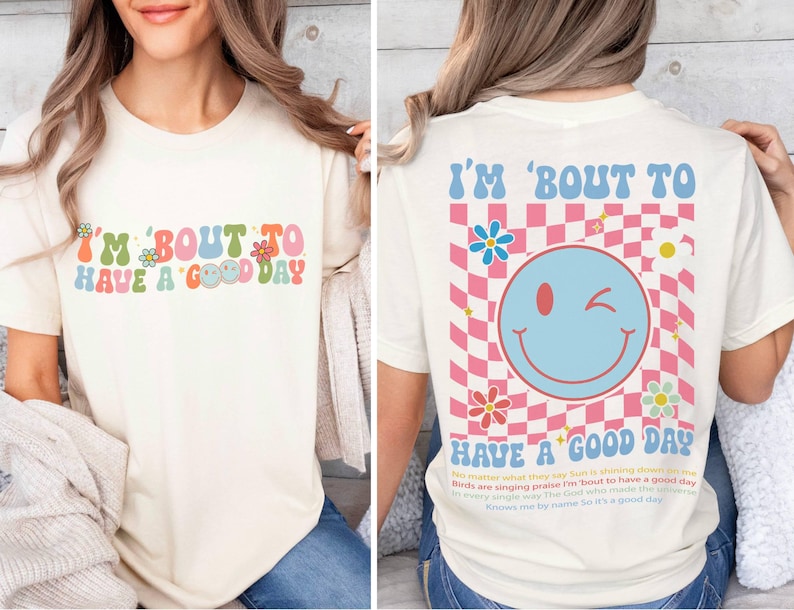 Puede incluir: Camiseta color crema con el texto "I'M 'BOUT TO HAVE A GOOD DAY" en letras coloridas de estilo retro. La parte trasera presenta una gran carita sonriente gui&ntilde;ando un ojo sobre un fondo a cuadros rosa y blanco, con flores y texto.