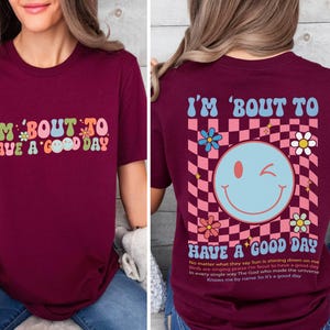 Puede incluir: Camiseta burdeos con el texto "I'M 'BOUT TO HAVE A GOOD DAY" en letras retro coloridas. La parte trasera presenta un patr&oacute;n de cuadros, una carita sonriente gui&ntilde;ando un ojo, flores y texto.