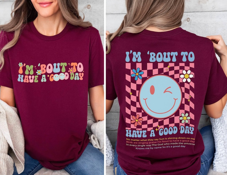 Puede incluir: Camiseta burdeos con el texto "I'M 'BOUT TO HAVE A GOOD DAY" en letras retro coloridas. La parte trasera presenta un patr&oacute;n de cuadros, una carita sonriente gui&ntilde;ando un ojo, flores y texto.